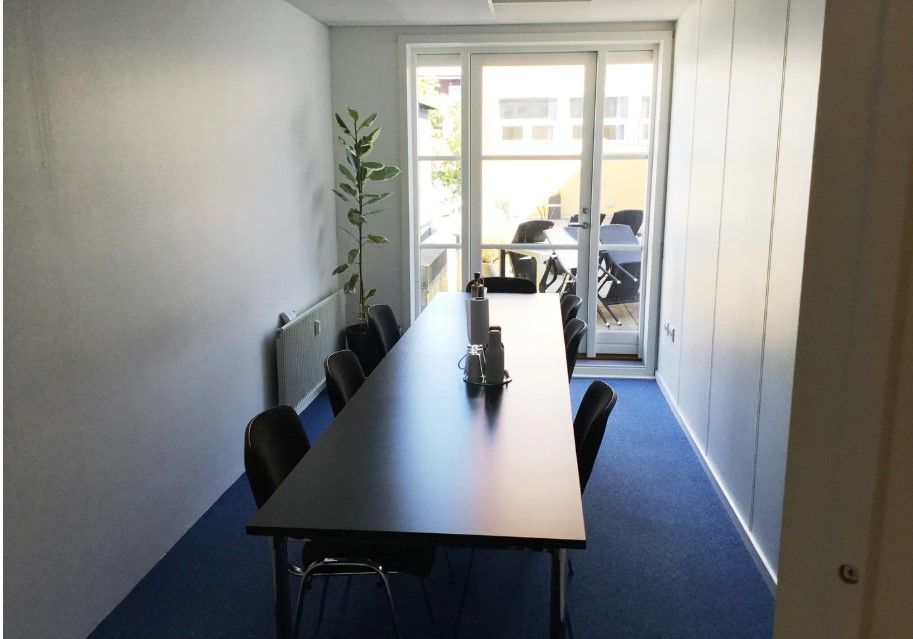 Kontorlokale med moderne møbler, en stor lang bord og lyseruder udenfor.