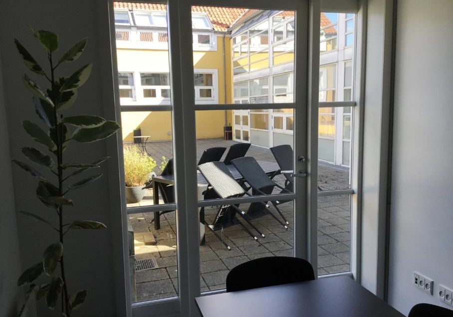 Kontorlokale med åbne ruder og bord med stålfødere i Aabenraa. Lyst kontor med udsigt til bygning og have med planter og