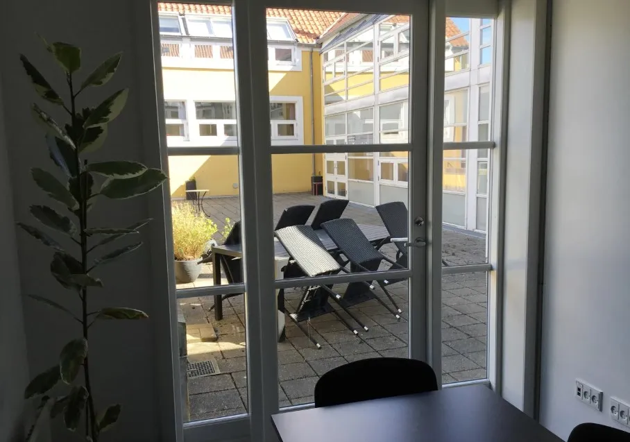 Kontorlokale med åbne ruder og bord med stålfødere i Aabenraa. Lyst kontor med udsigt til bygning og have med planter og