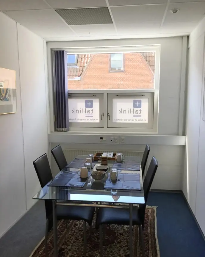 Kontor med en stor glasbord og fire stole samt en vinduesvisning af en murstensbygning med træloft og vindue