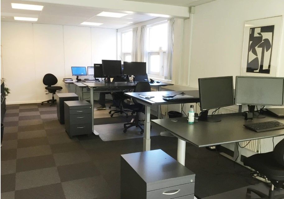 Moderne kontorlokale med lyse loftslamper, flere skriveborde, stolpe og computere samt en vægmaleri og en plante