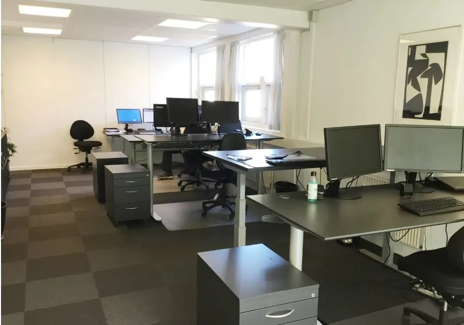Moderne kontorlokale med lyse loftslamper, flere skriveborde, stolpe og computere samt en vægmaleri og en plante