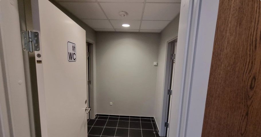 Kort gang med hvide døre og mørke gulvfliser. Der er et WC-skilt med et mand og kvinde ikon. Der er en indbyggede lampe og