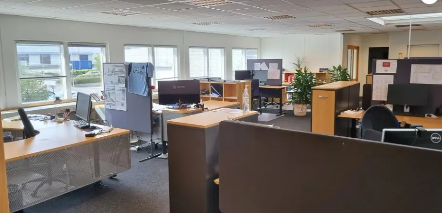 Kontor med lyse gulve, høje ruder og moderne møbler, herunder computere og skriveborde. Mange plantager og lyse kontorer.