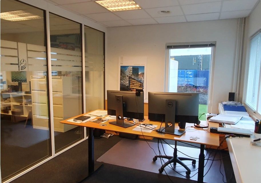 Kontor med to Dell-skærme, skrivebord, stol, kabinet, postkasse, glasvinduer og udgang til gaden