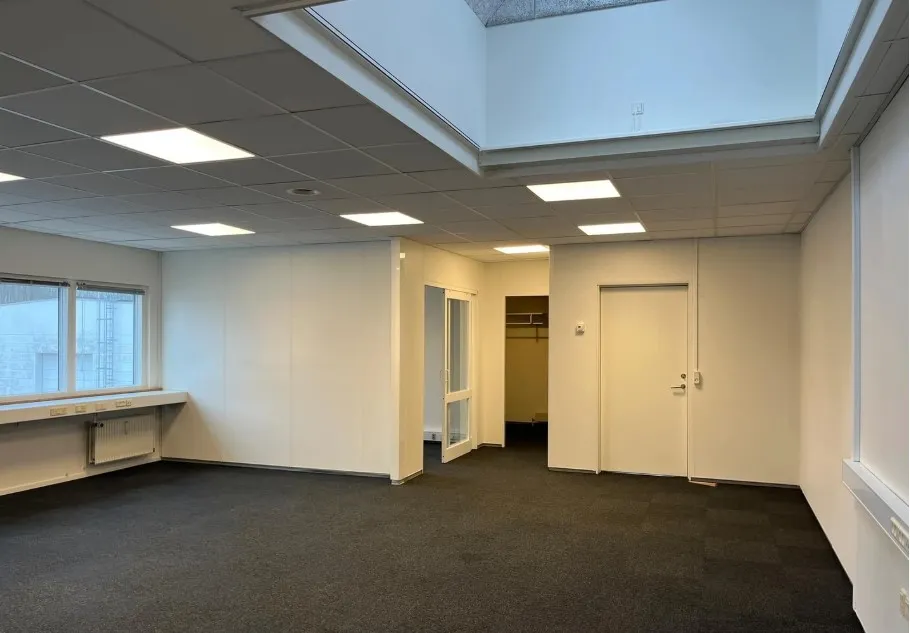 En stor, tomt kontor med lyseramme og hvide vægge samt en åben dør og et vindue med udsigt til bygningen og trappen.