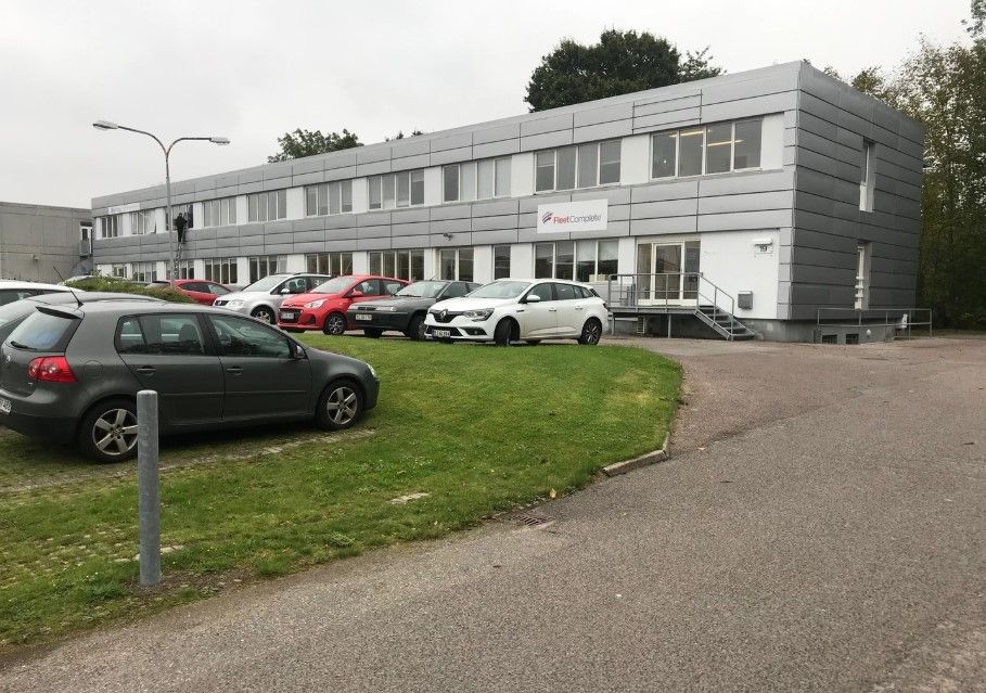 En stor hvid og grå bygning med flere biler parkeret udenfor og et skilt med teksten Fleet Complete