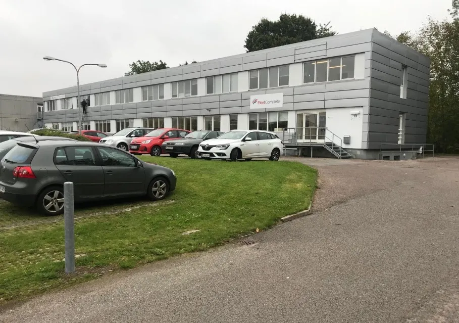 En stor hvid og grå bygning med flere biler parkeret udenfor og et skilt med teksten Fleet Complete