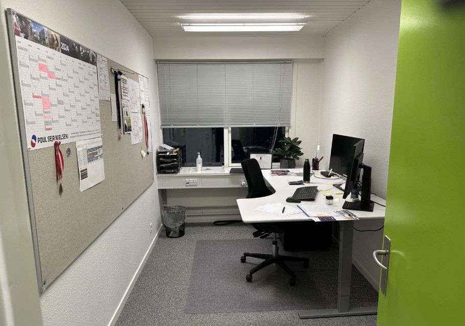 Plakater med Poul Seier Nielsen 2024, en stol, en skrivebord, en computer, et tastatur, en plante og en papirkurv i et