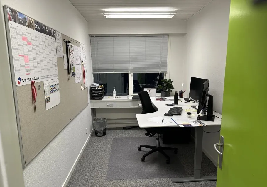 Plakater med Poul Seier Nielsen 2024, en stol, en skrivebord, en computer, et tastatur, en plante og en papirkurv i et