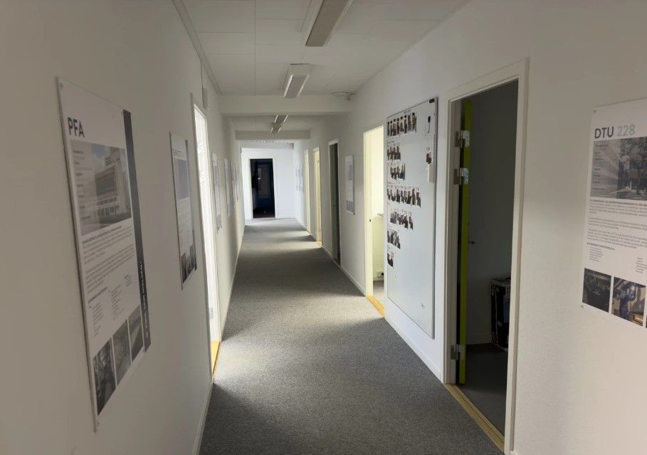 Lang korridor med hvide vægge, gulvbelægning og vægpapir med billeder og tekst, samt åbne døre og døre med håndtag.