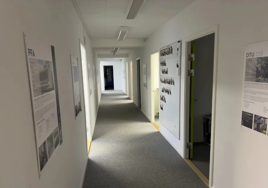 Lang korridor med hvide vægge, gulvbelægning og vægpapir med billeder og tekst, samt åbne døre og døre med håndtag.