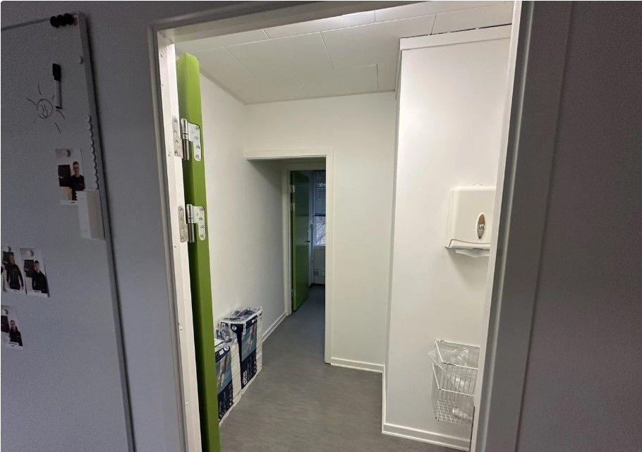 Kort gang med grønne døre og hvide vægge. På højre side hænger en hvid papkasse. På væggen til venstre er der et magnetbræt