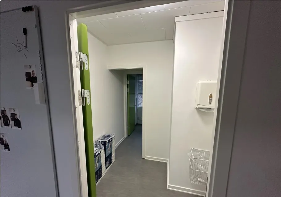 Kort gang med grønne døre og hvide vægge. På højre side hænger en hvid papkasse. På væggen til venstre er der et magnetbræt