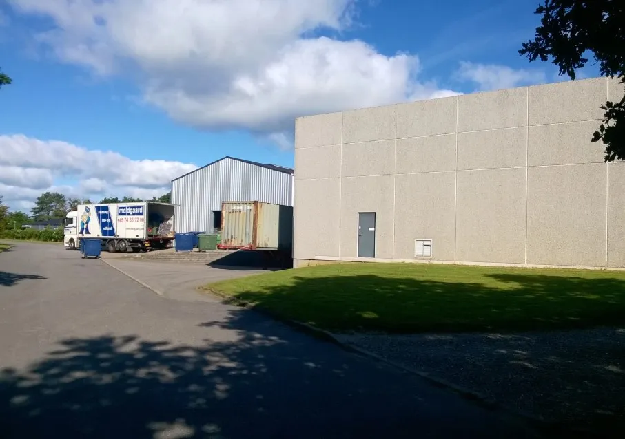 Forretningslokaler med parkeringsplads og lastbil ved siden af i Løsning. Foran er en grøn buskethæk og skrapelukket gulvej