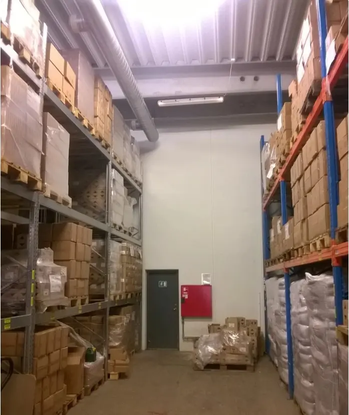 Lager med varer og sager, lyse og hvide vægge, indgangsdør med sikkerhedsskilt og brandvæsen, røde døre og paller med sække