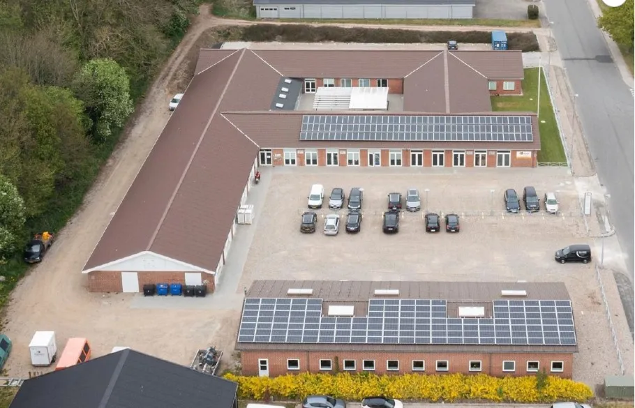 Kontorbysnit med solpaneler på taget og flere parkerede biler i parkeringspladsen. Solpanelerne sikrer bæredygtig energi, og
