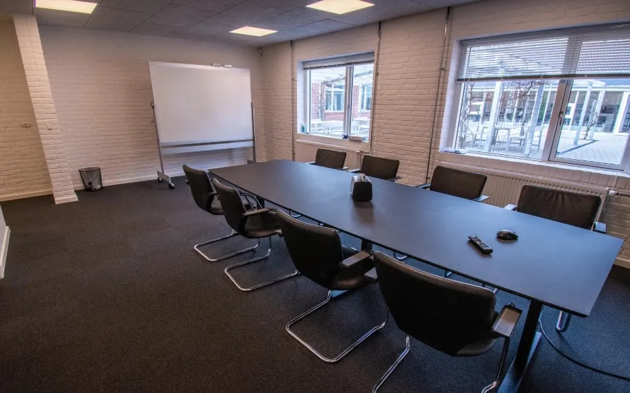 Kontor med langt bord, stolpe, projektor og vindue med udsigt til bygning i baggrunden