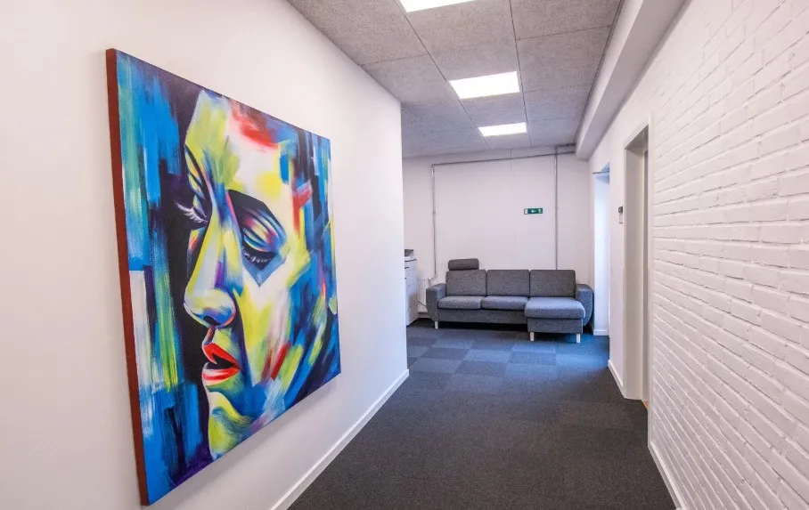 En stor farverig maleri af et kvindeansigt er hængt på væggen i en kontorgang. En sofa er stillet op langs væggen og der er