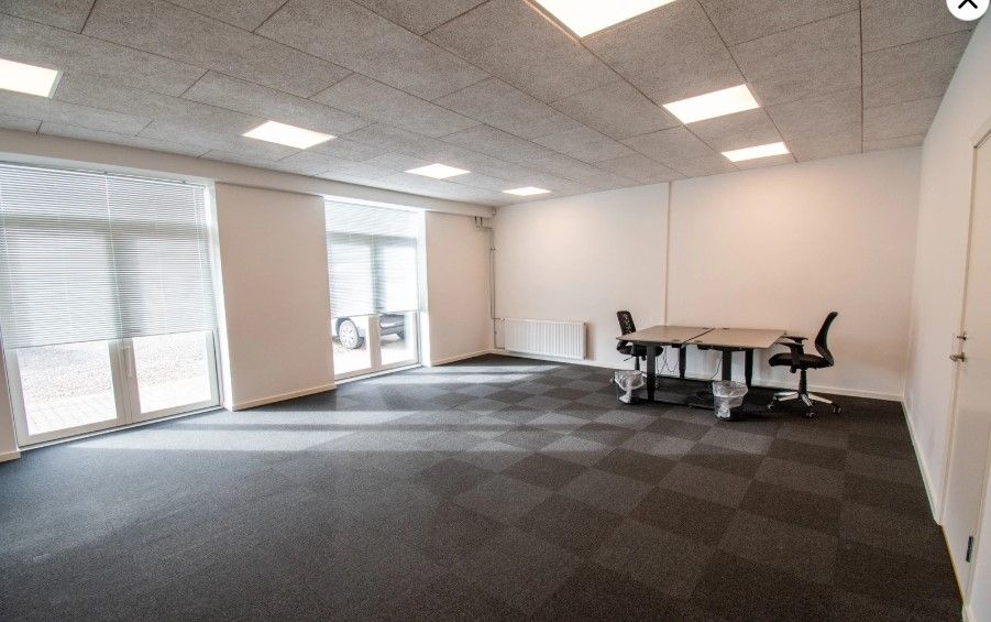 Et tomt kontor med to skriveborde, to stole og to skraldespande. Væggene er hvide, og gulvet er grønt og hvidt. Der er