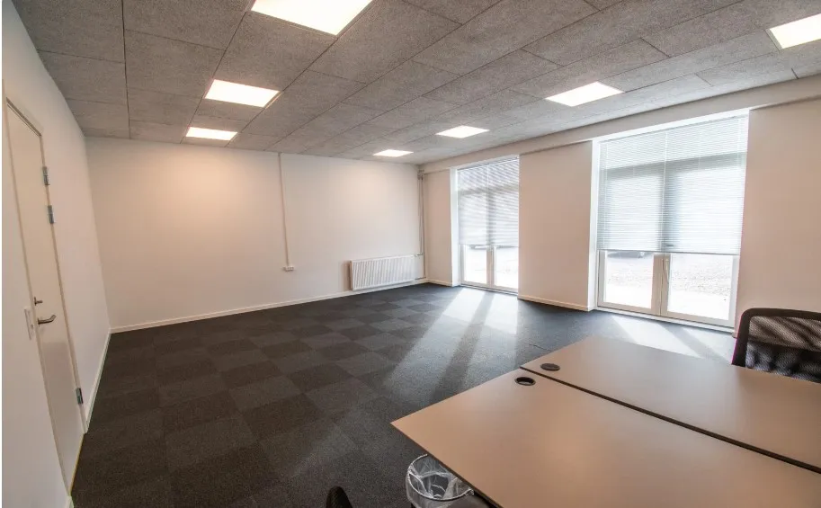 Kontor med hvide vægge, karnapper, to bordsæt og to vinduer med gardiner samt en række indbyggede lys i loftet.