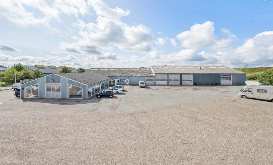 En stor industribygning med garagedør og parkeringsplads, hvor der er parkeret biler og en rv