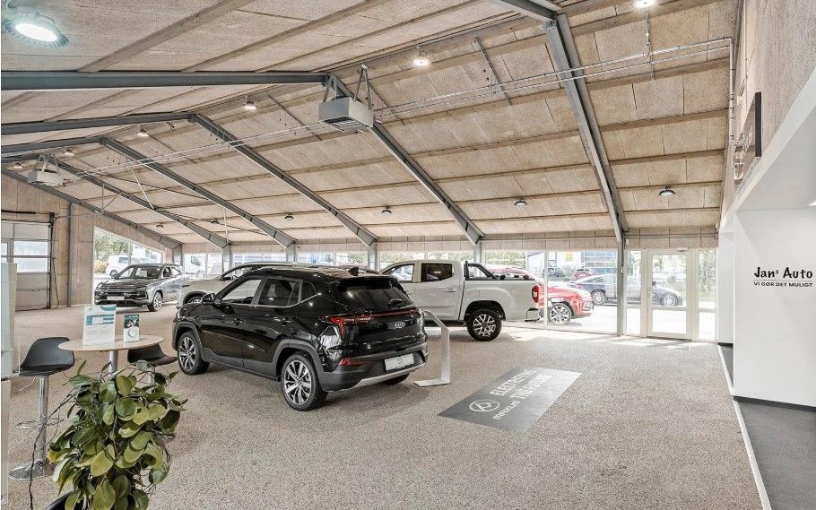 I en industribygning står flere biler udstillet, herunder en sort SUV, en hvid pickup, og en gul bil. Der er også en grøn