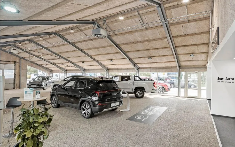 I en industribygning står flere biler udstillet, herunder en sort SUV, en hvid pickup, og en gul bil. Der er også en grøn