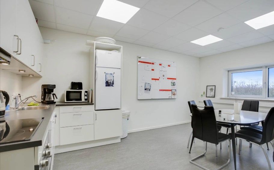 Køkken med lyse loftslampe og hvide vægge samt køleskab, mikrobølgeovn og bord med stole