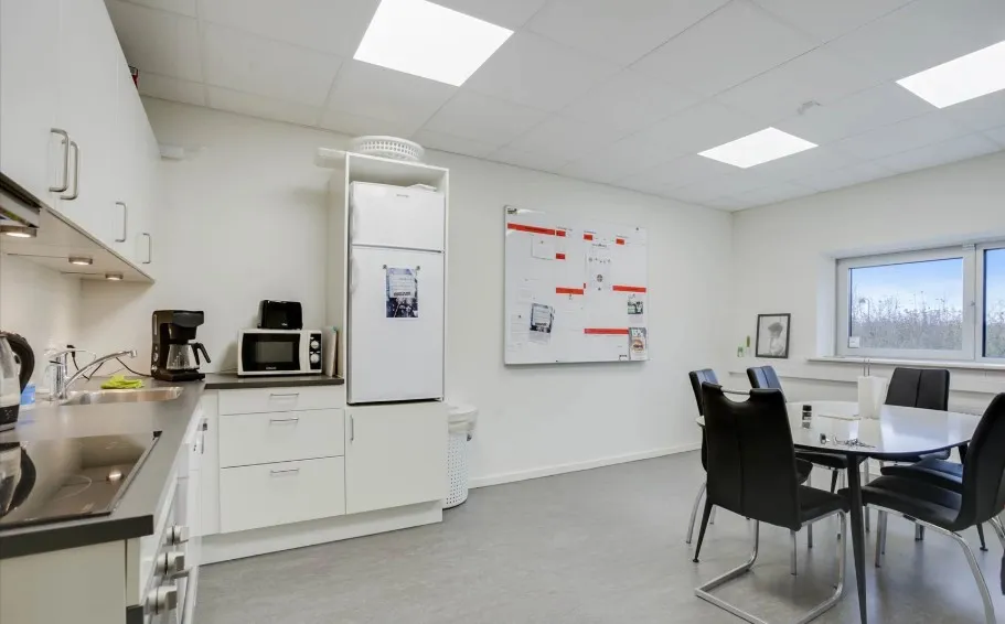 Køkken med lyse loftslampe og hvide vægge samt køleskab, mikrobølgeovn og bord med stole