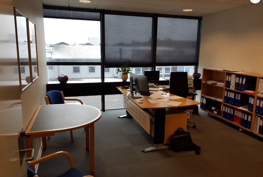 Kontor med vinduer, skrivebord, computer og en vase med blomster, stol og hylde med papirer og filer.