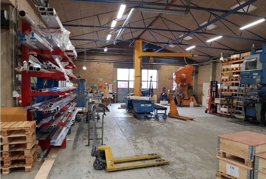 Stort produktionslager med flere kranarme, forskellige lagerreoler, møbler og apparater, samt træpæle og en gul gaffeltruck.