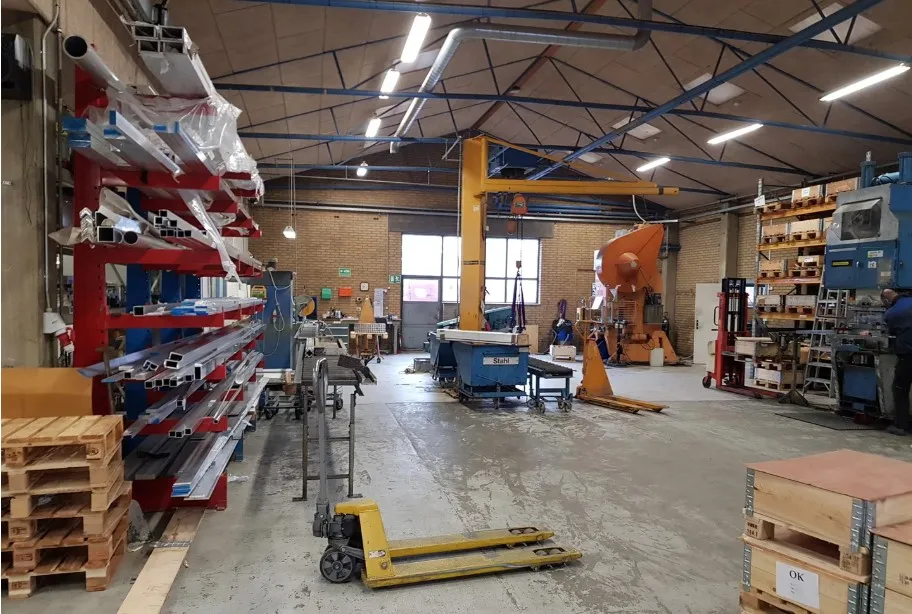 Stort produktionslager med flere kranarme, forskellige lagerreoler, møbler og apparater, samt træpæle og en gul gaffeltruck.