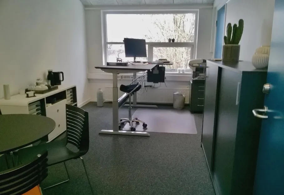 Kontor med computer, skrivebord, stol, kaffemaskine, vasketøj, radiator, og plante samt en dør