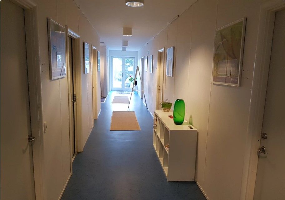 Korridor med lysegul væg og gulgul gulv med flere døre og en hvid skuffe med grøn vase og plante samt lysekroner og malerier