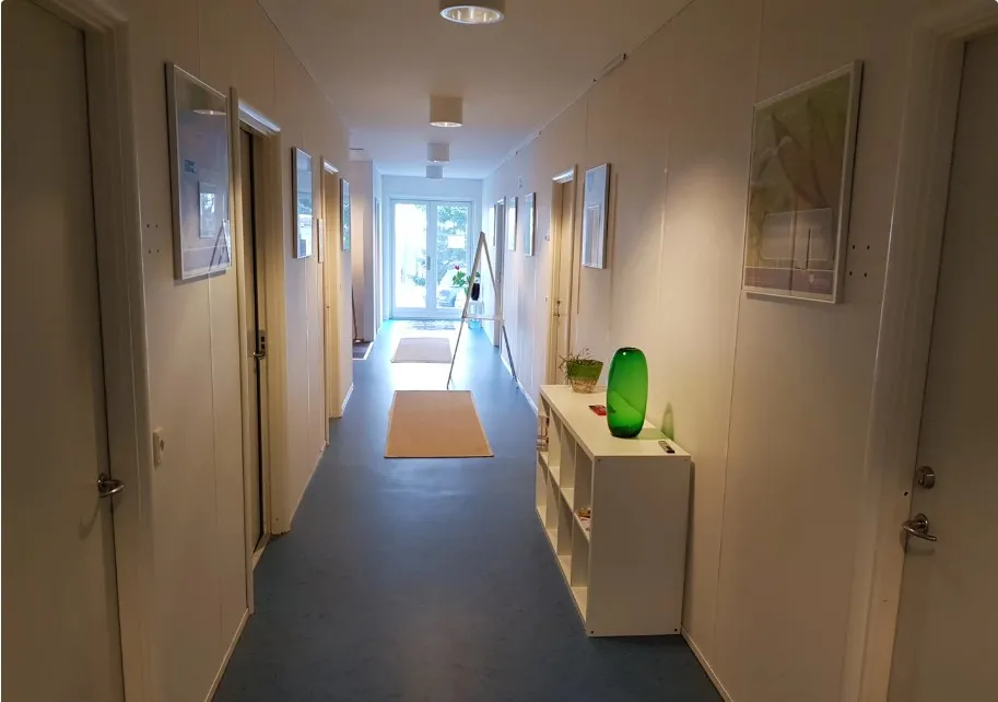 Korridor med lysegul væg og gulgul gulv med flere døre og en hvid skuffe med grøn vase og plante samt lysekroner og malerier