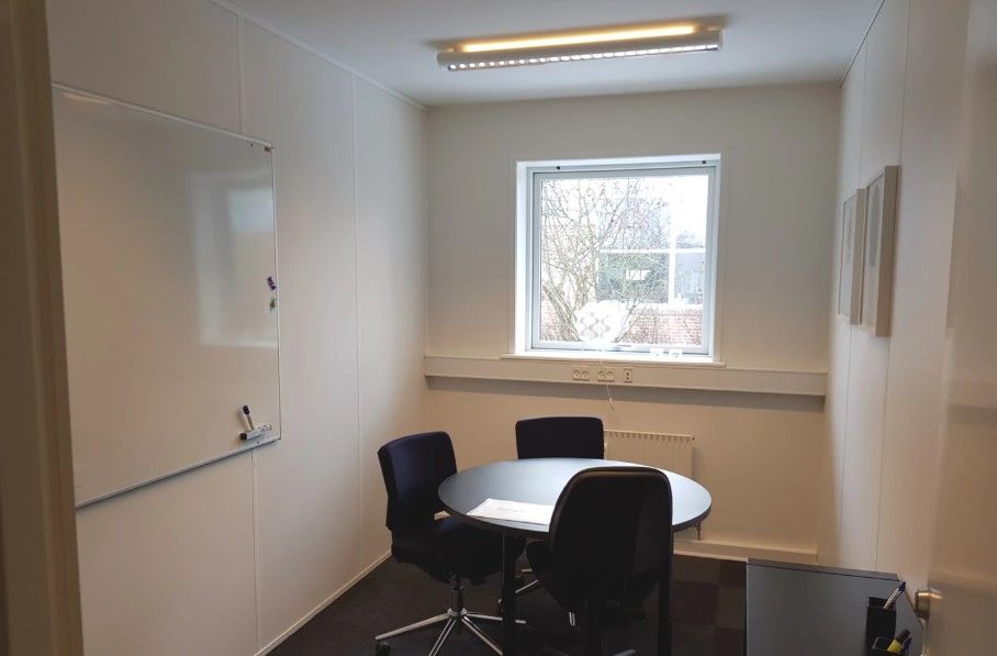 Kontor med runde bord, stol, whiteboard, radiator, vinduer, lysekroner og lysegulv