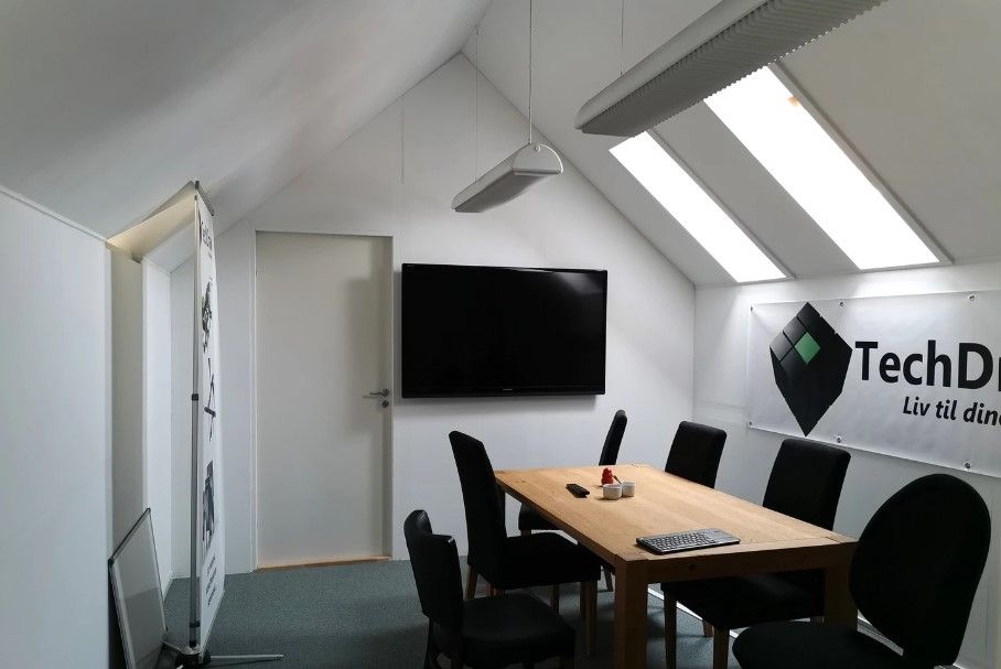 En mødelokale med skrivebord, stole og en tv-skærm, samt et banner med TechD.