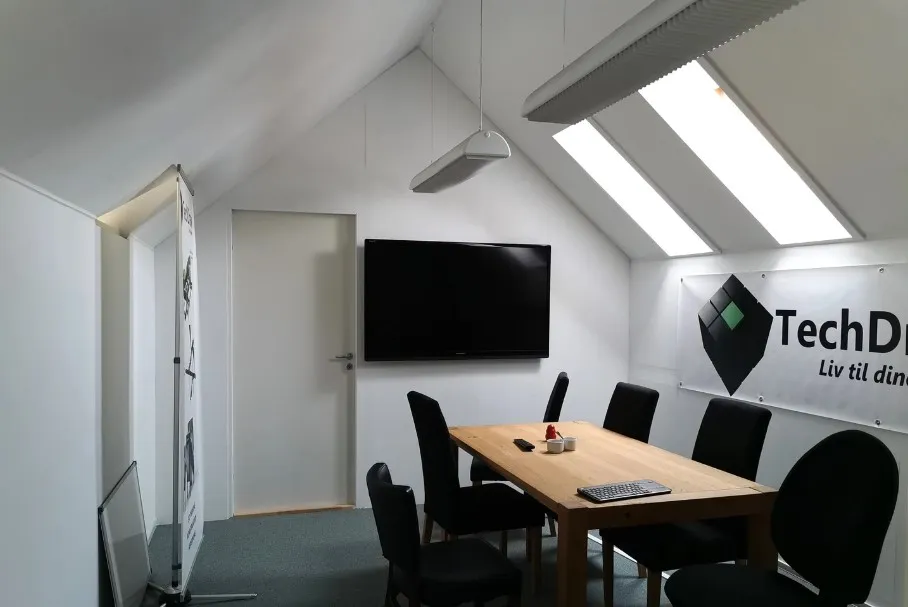 En mødelokale med skrivebord, stole og en tv-skærm, samt et banner med TechD.