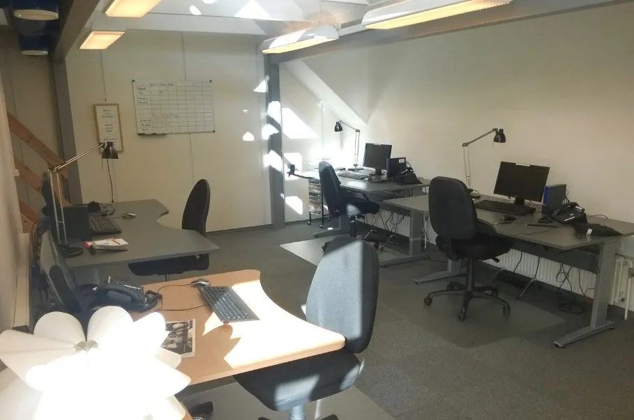 Kontorrum med tre skriveborde, hver med computer og skrivebordslampe. Væggene er hvide og der er et tavle med noter og en