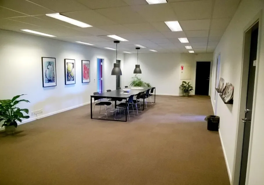 Kontor med langt bord og stole i midten og tre billeder på væggen. En plante og en skraldespand til højre.