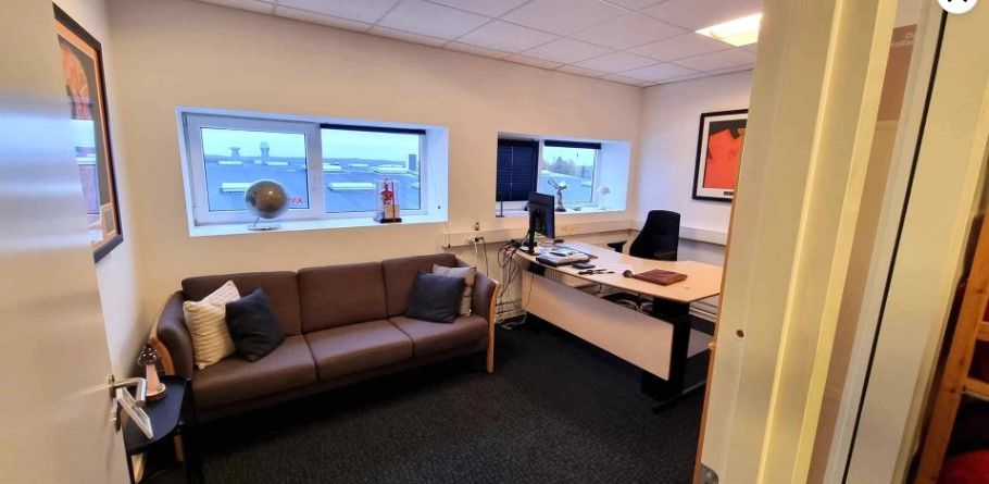 Et kontor med sofa, bord, stol og computer, og to vinduer med udsigt til byen og rødt maleri på væggen