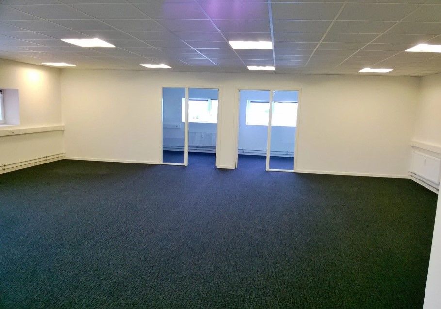 Et tomt kontor med hvide vægge, glasdøre og glasruder samt lyskappe på loftet. Gulvet er dækket af grønt gulvtæppe.
