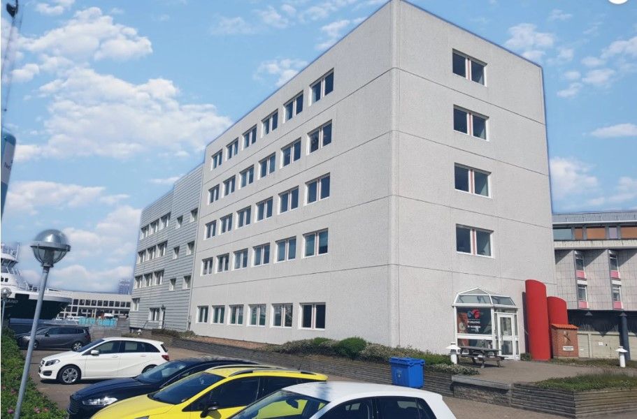 et kontorbygning med lysegrå facade og mange vinduer i Esbjerg. Der er parkeret biler udenfor, og der er også en rødgule
