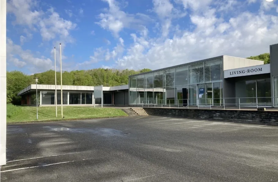 En stor moderne bygning med store glasruder og en stor parkeringsplads foran. Der er en græsplæne og nogle trær og skyer på