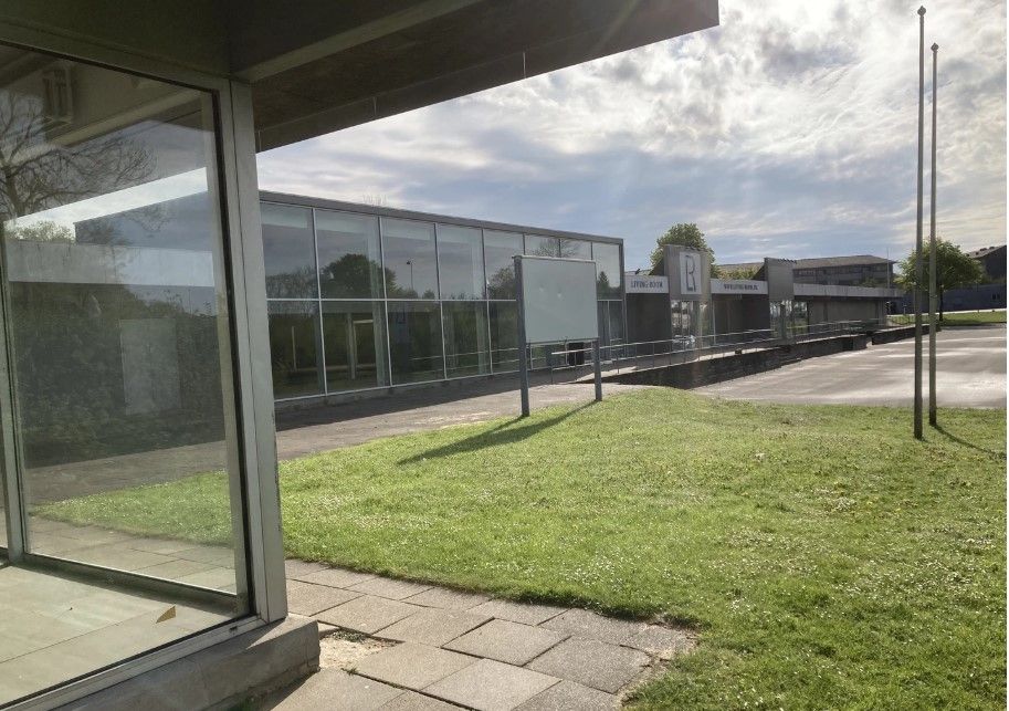 Glasdøre og -ruder i moderne bygning, udendørs græsplæne og solrig himmel med skyer, to slanke flaggestænger og træer i