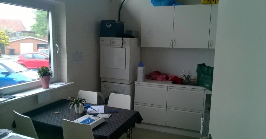 Et køkken med hvide vægge og møbler og en lille stue med skrivebord og blomster.