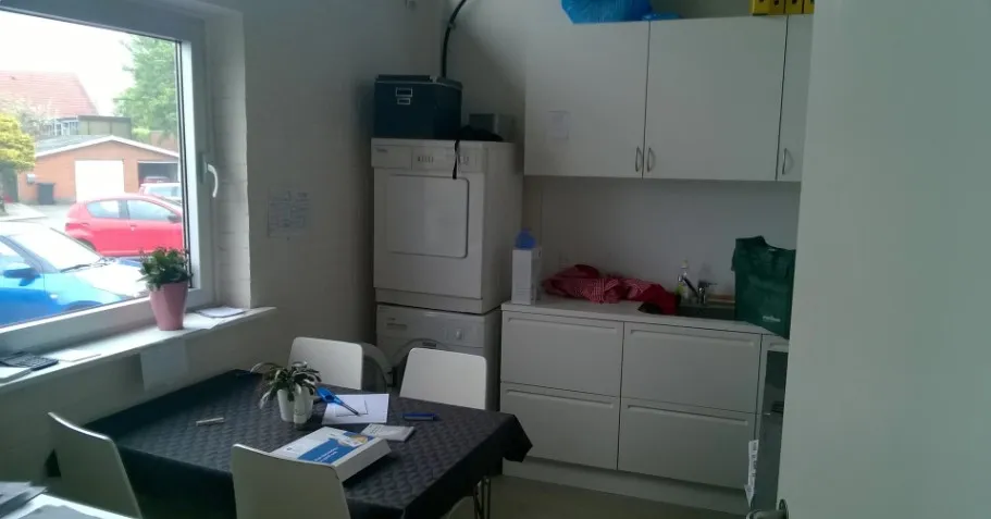 Et køkken med hvide vægge og møbler og en lille stue med skrivebord og blomster.