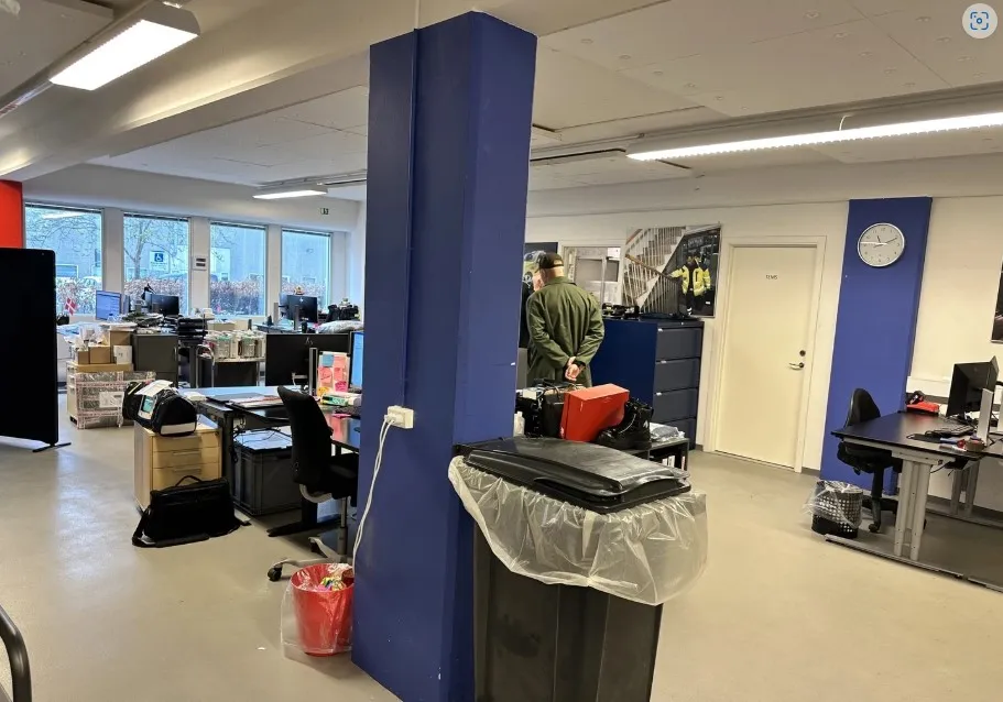 Kontor med flere bord, stol, og computere samt en stor papkasse og en høj blå søjle