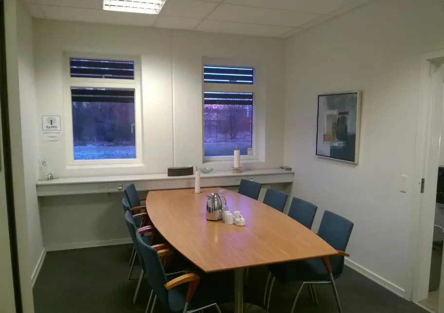 En tom møde- og konferencesal med et brun træbord og flere blå stole, der er placeret rundt om bordet